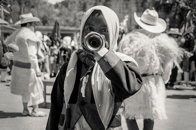 Un niño músico vestido con su traje tradicional tzotzil y un pañuelo blanco envuelto en la cabeza, toca una trompeta corta. Ve a cámara con mirada tierna. Al fondo hay dos hombres vestidos con taje de gala de borrego blanco, con sombreros y más al fondo, está la comunidad, tocando y bailando en las montañas.